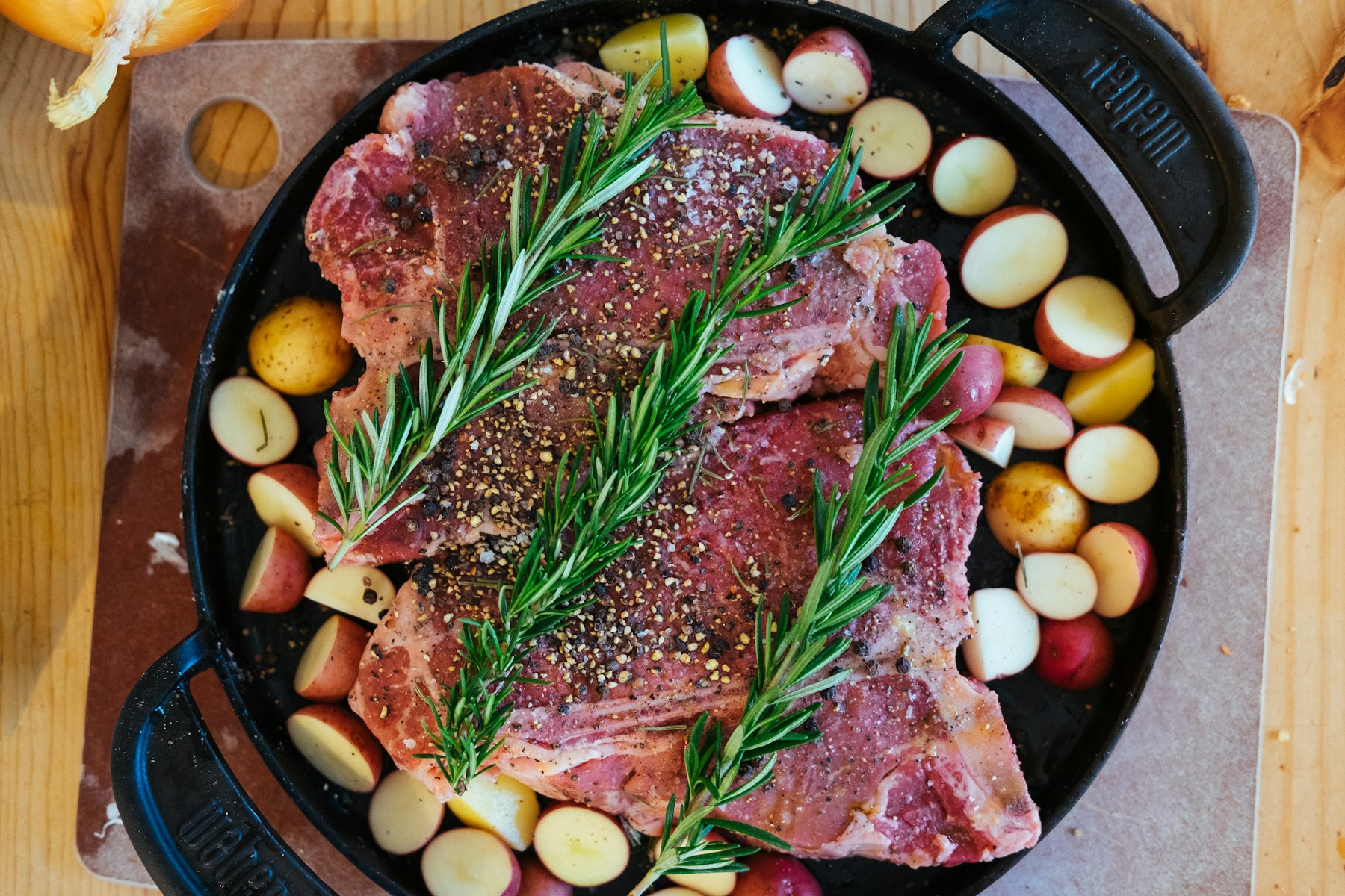 Seasoned raw steaks with rosemary and potatoes in a cast-iron pan