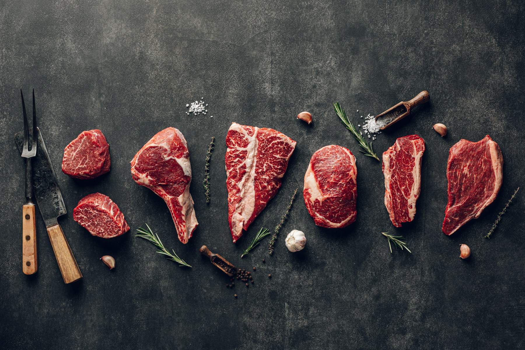 Assorted raw steak cuts arranged with knives, garlic and herbs