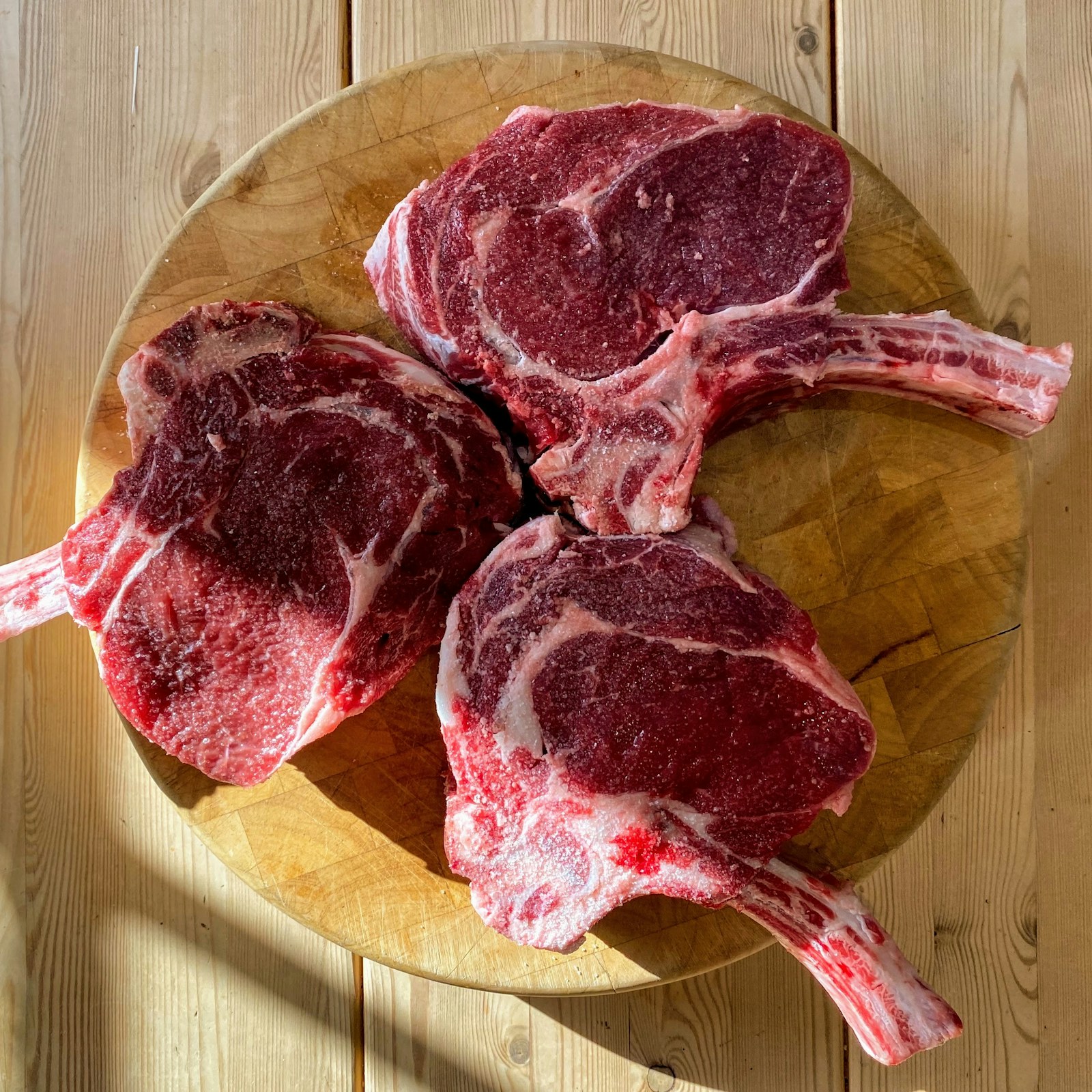 Three thick raw bone-in ribeye steaks on a wooden board